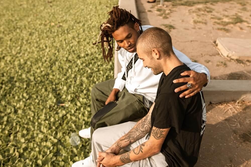 A young man sitting with his arm around a friend who is looking down.