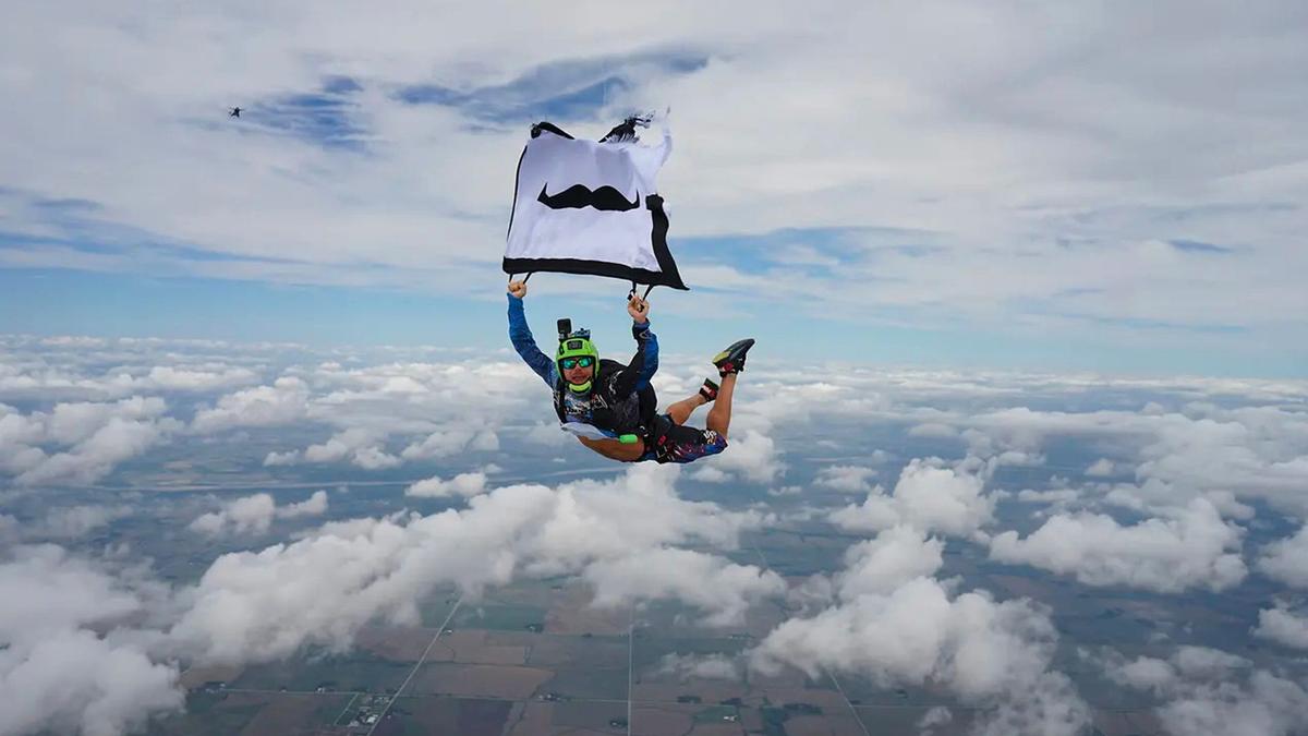 A man skydiving holding up a white flag with a mustache imprinted on it.