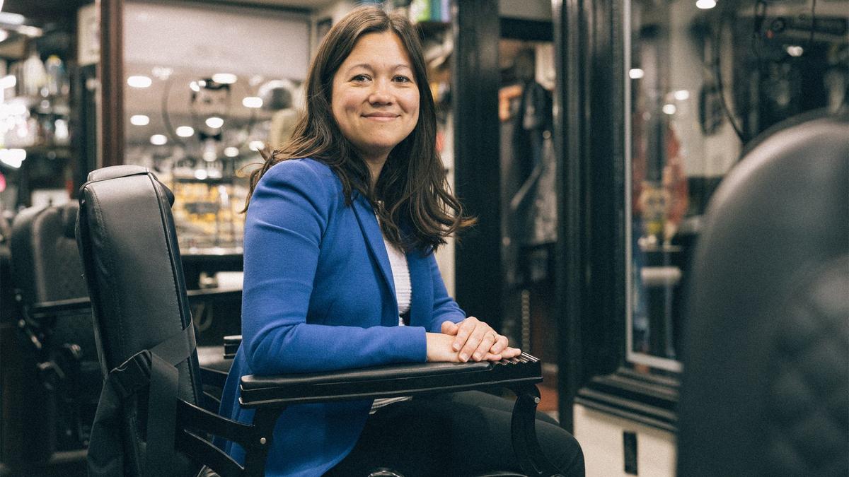 A woman in a blue jacket poses for a portrait from a barber chair in a barber shop