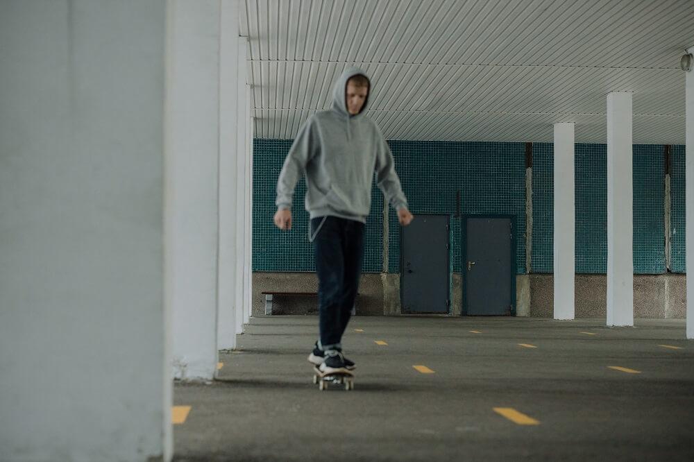 A young man in a grey hoodie skateboards alone in an empty undercover car park.