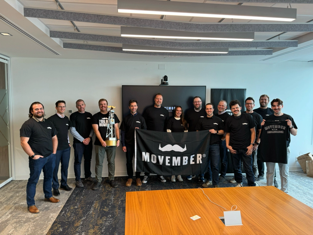 A group of Movember Ambassadors are standing together wearing Movember merch and smiling at the camera