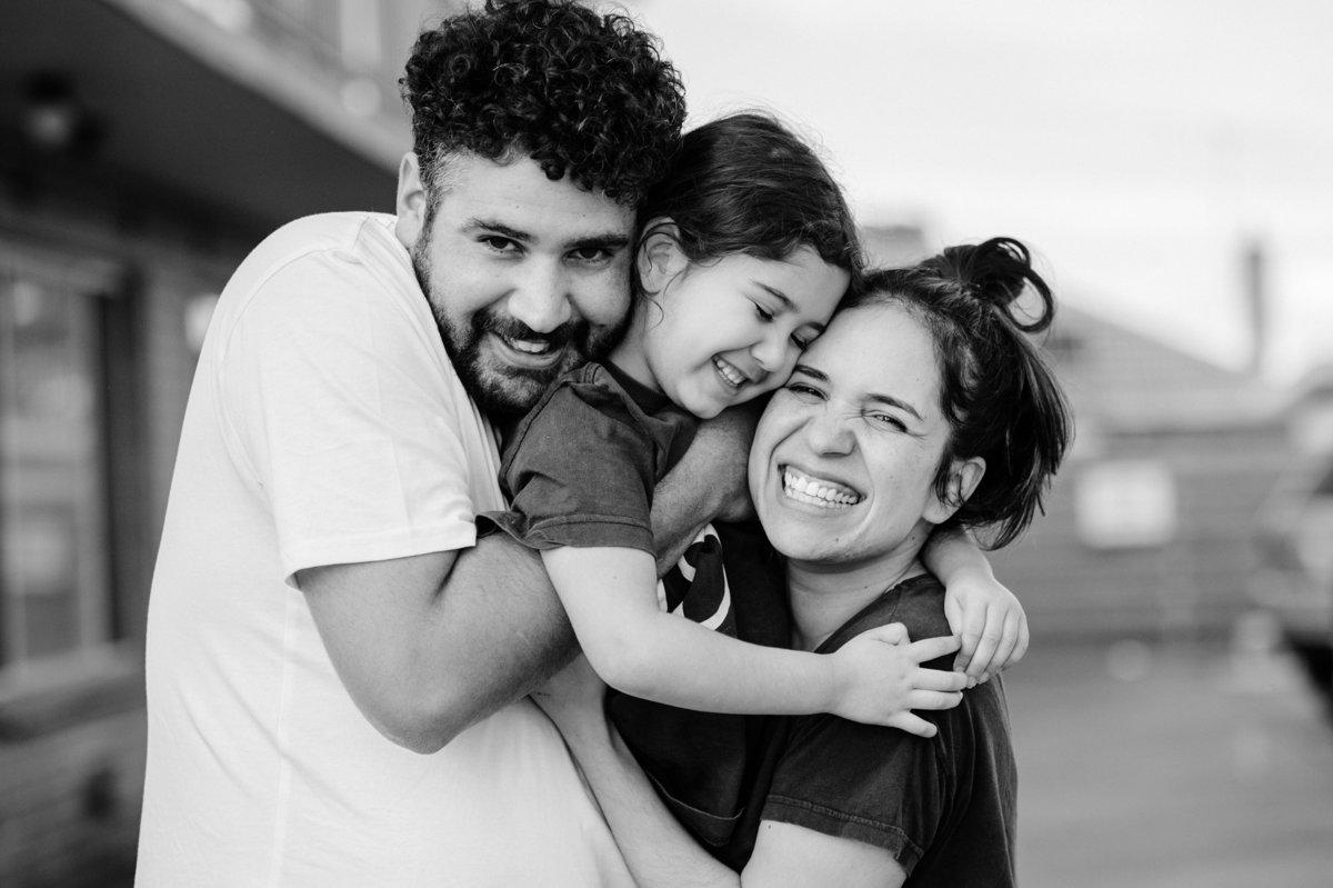 A young child hugs and laughs with both parents outdoors.