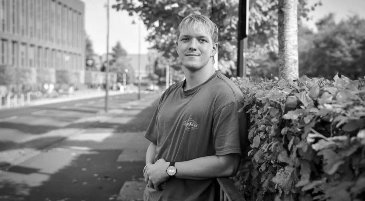 Man standing on a street smiling and leaning against a hedge