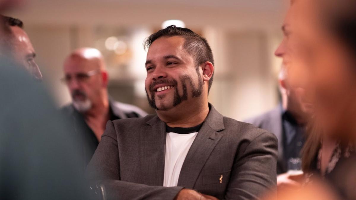 Man with unique shaved moustache design, in suit, stands with others around him at event