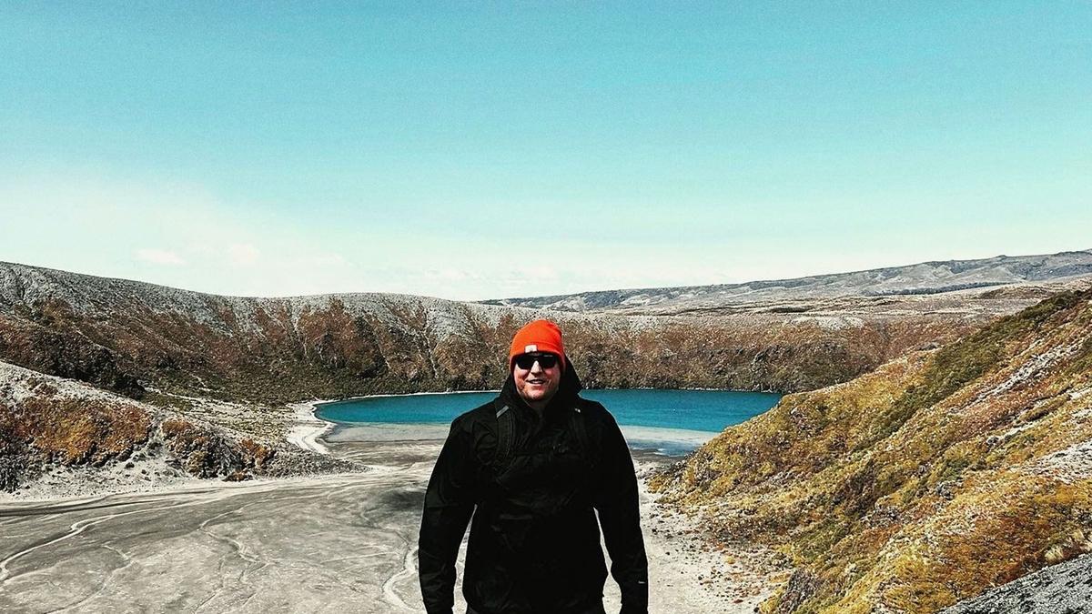 Phil stands on top of a mountain with a majestic blue lake behind him. He wears winter clothes for the mountain conditions.