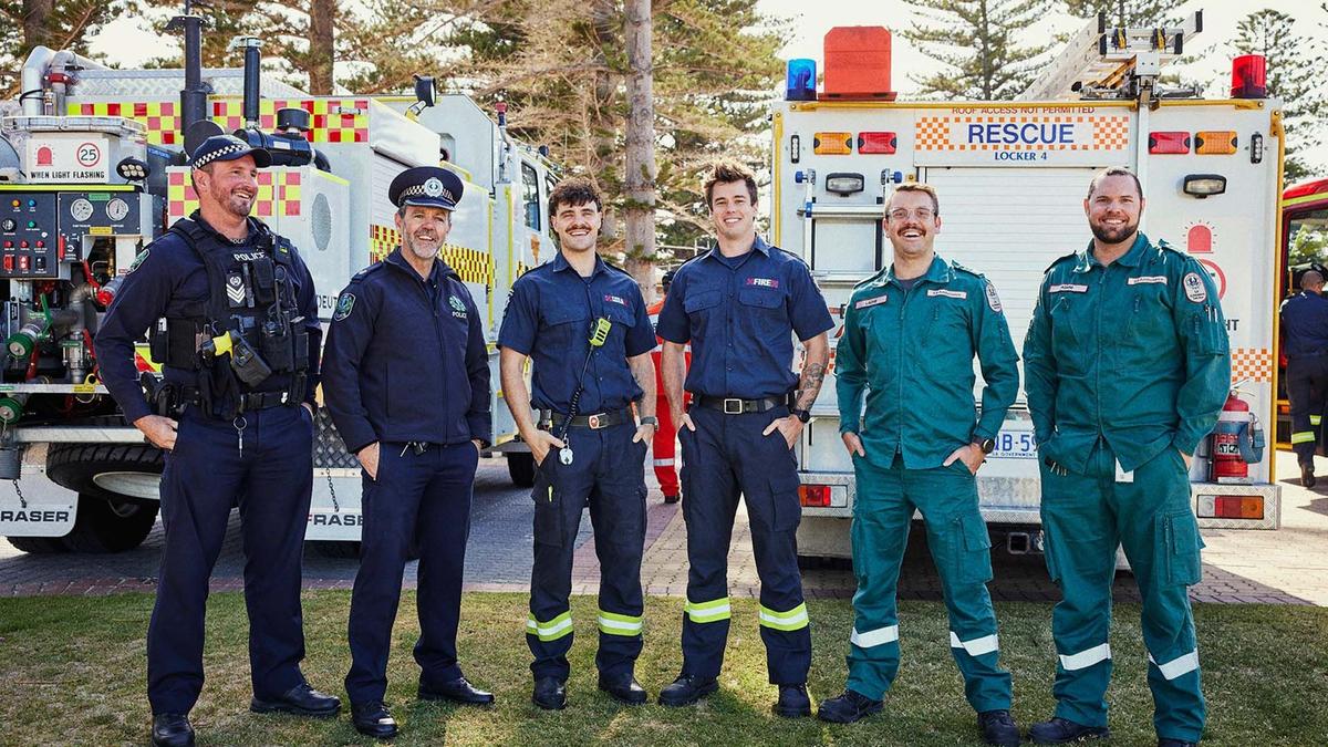 A group of emergency services men stand in a line, all in uniform, with their emergency vehicles behind them.