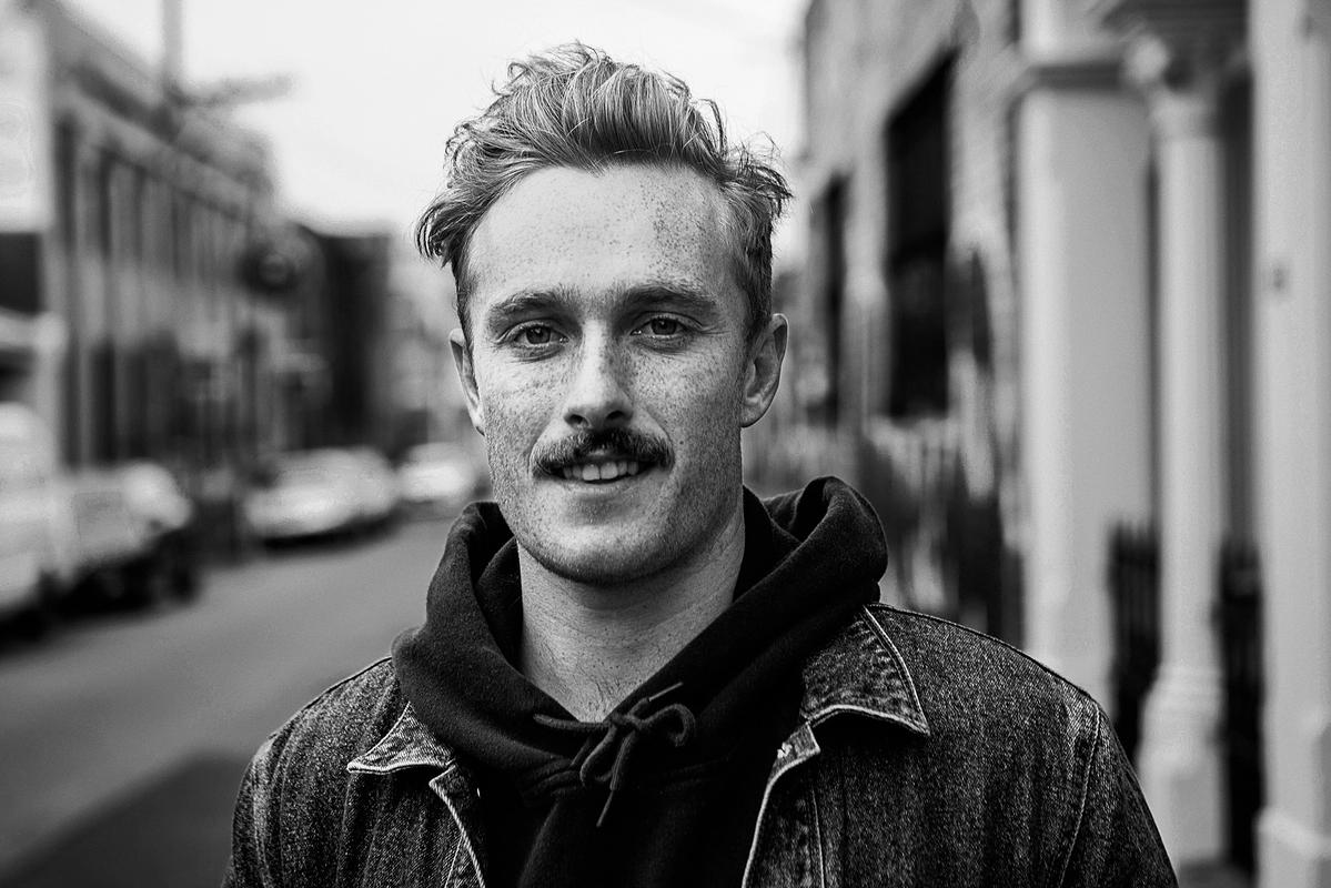 A black and white photo of a man with a moustache smiling slightly and looking directly at the camera