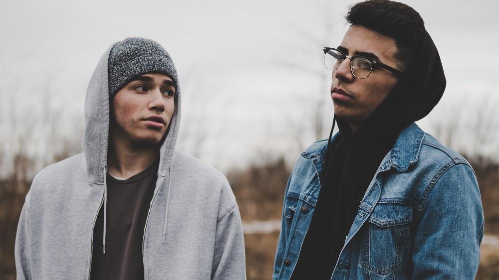 Two young men standing outdoors, one glancing over at his friend with a concerned expression.