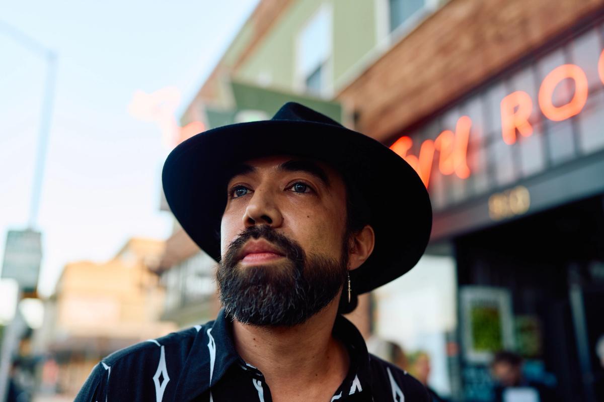 A man wearing a hat is looking off to the distance, we can see the neon sign of the Ivy Room behind him.