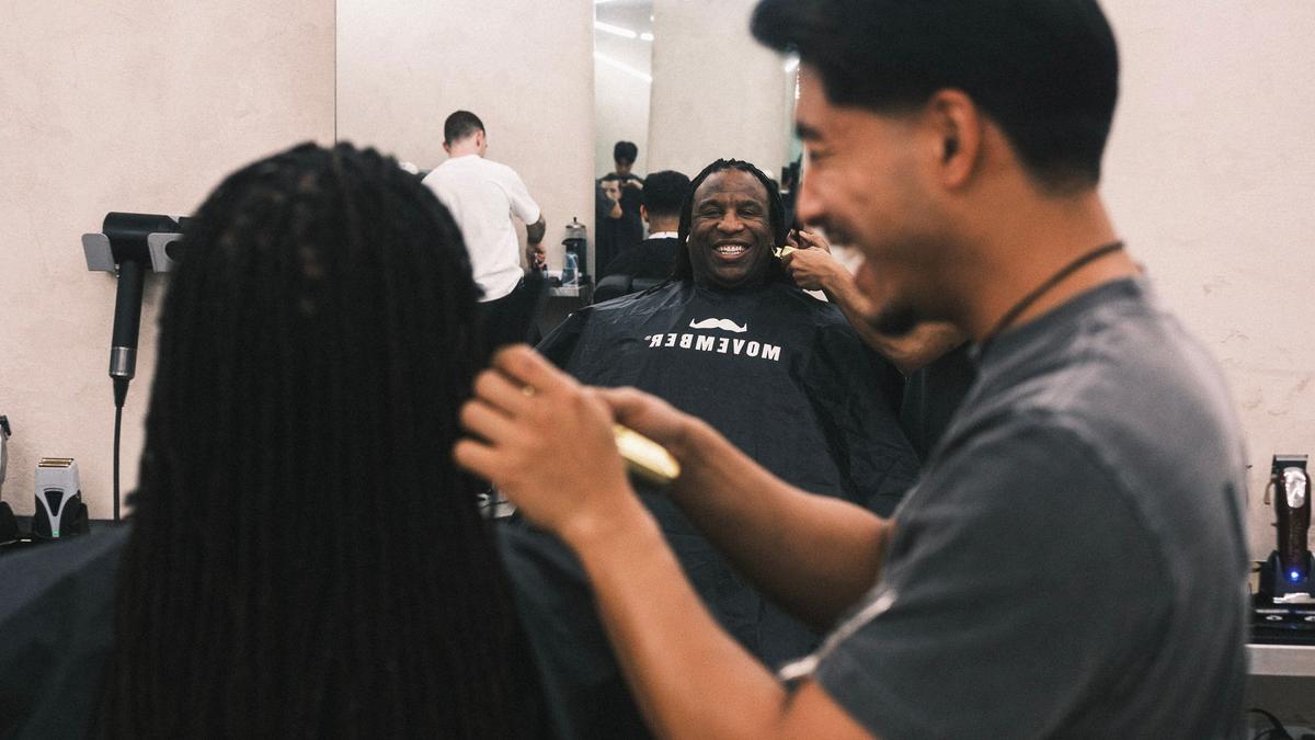 Photo taken of a man getting a shavedown, we see the bak of his head and the barber in the foreground, and his smiling reflection in the mirror