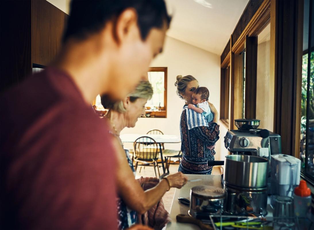 A mother is holding her baby in the kitchen while the father and grandmother can be seen in the foreground preparing a meal