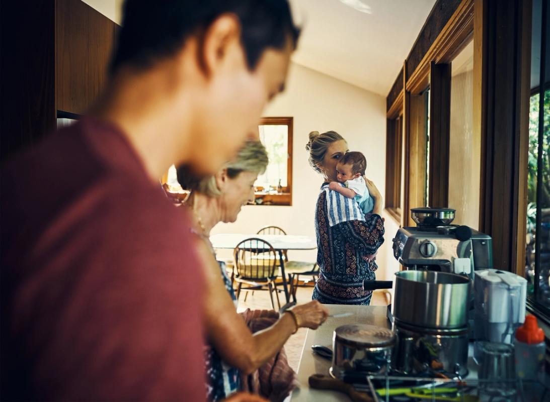 A mother is holding her baby in the kitchen while the father and grandmother can be seen in the foreground preparing a meal