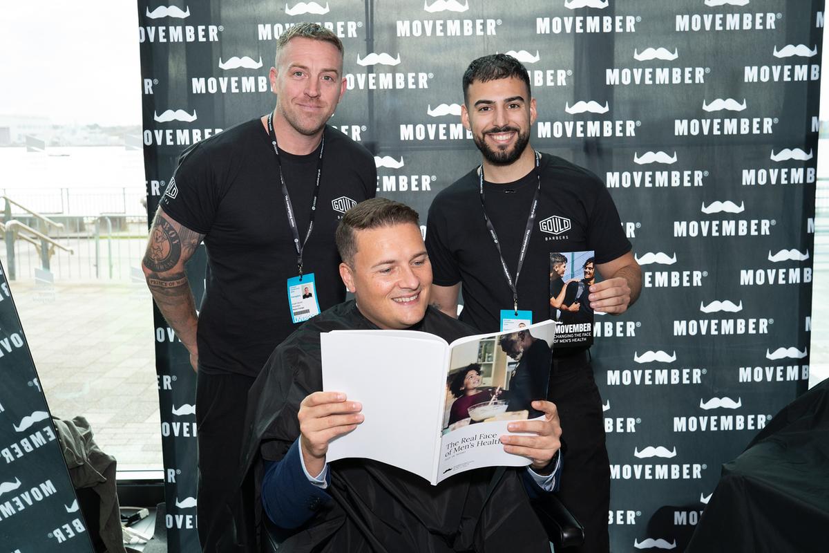 A man is sitting in a barber chair reading Movember's Real Face report, while two barbers stand smiling behind him
