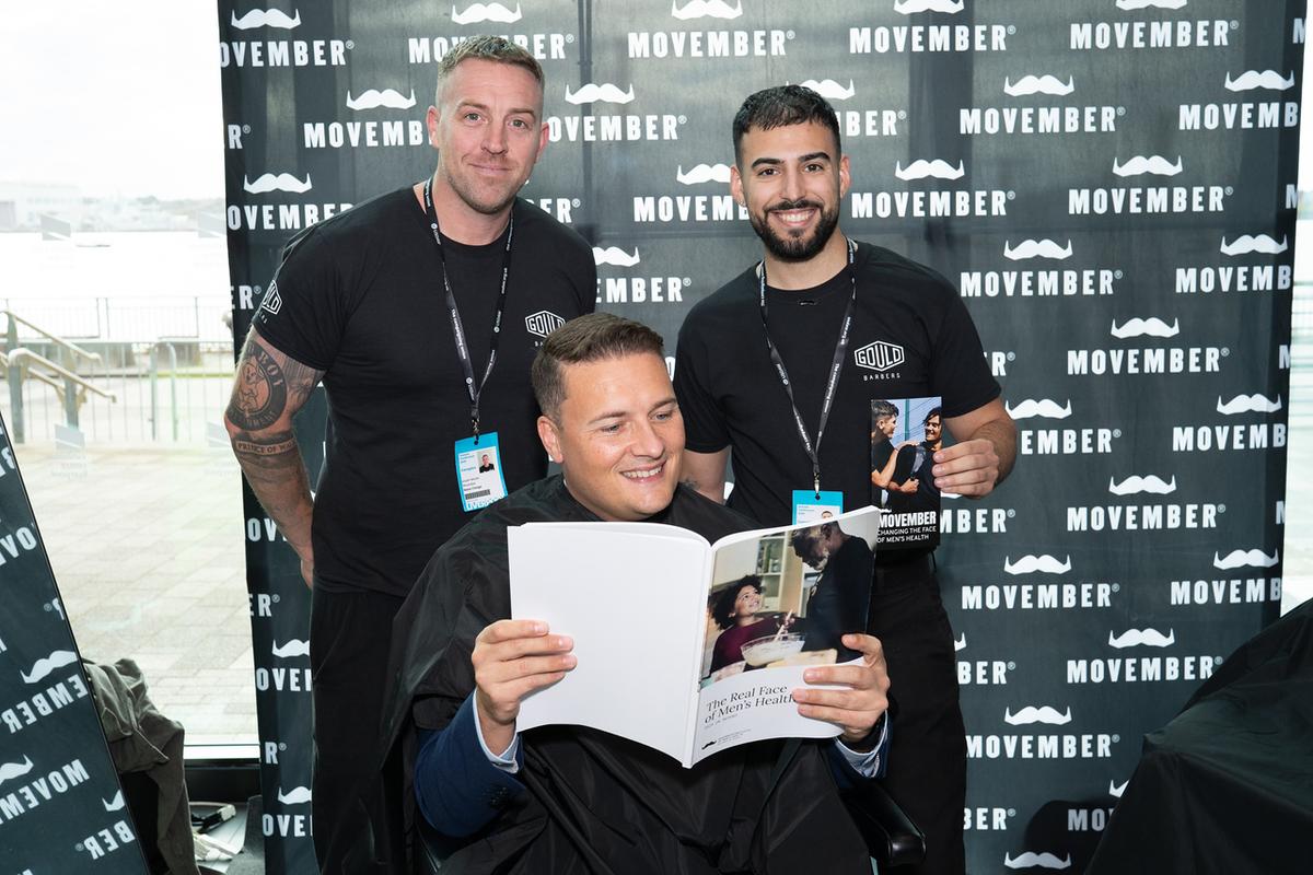 A man is sitting in a barber chair reading Movember's Real Face report, while two barbers stand smiling behind him