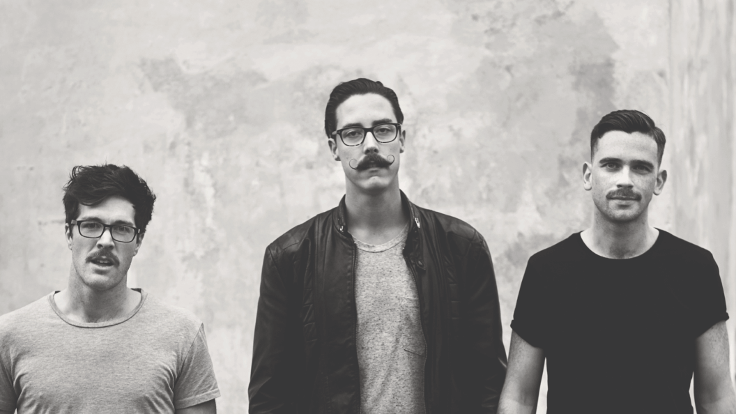 Three young men with moustaches stand side by side against a concrete wall, looking directly at the camera, in a black and white portrait.