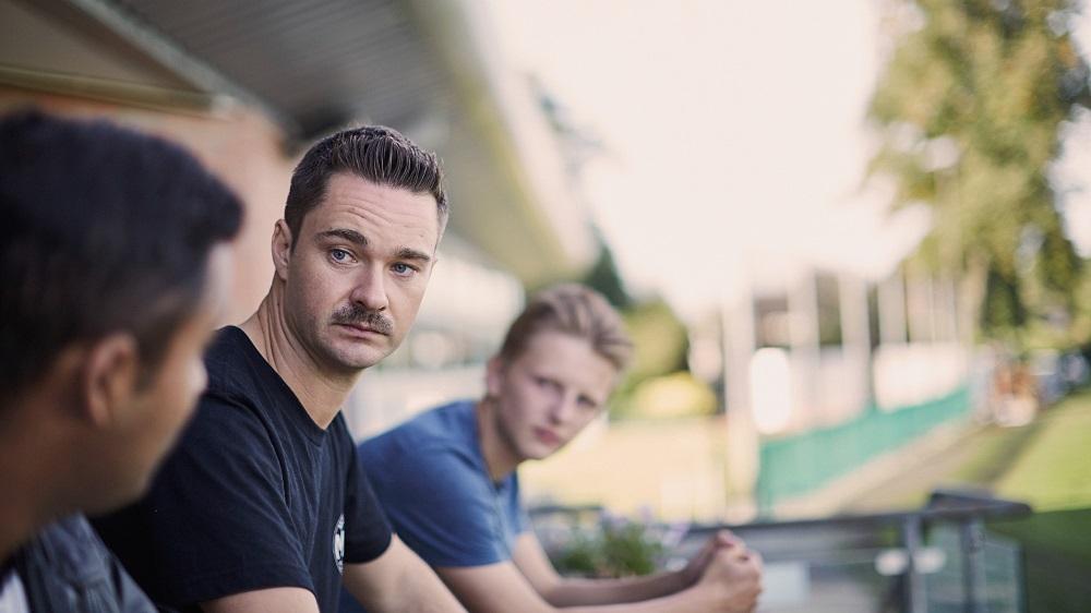 Three men sit together outdoors in conversation, one man looking directly at the camera with a serious expression.