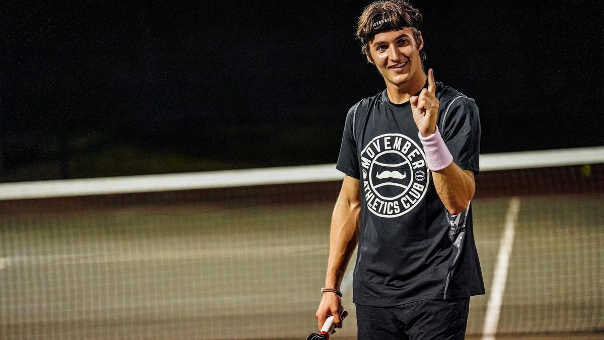A pickleball player is mid-game on the court, sporting a Movember t-shirt and sweatband, and pointing a Number 1 with his finger