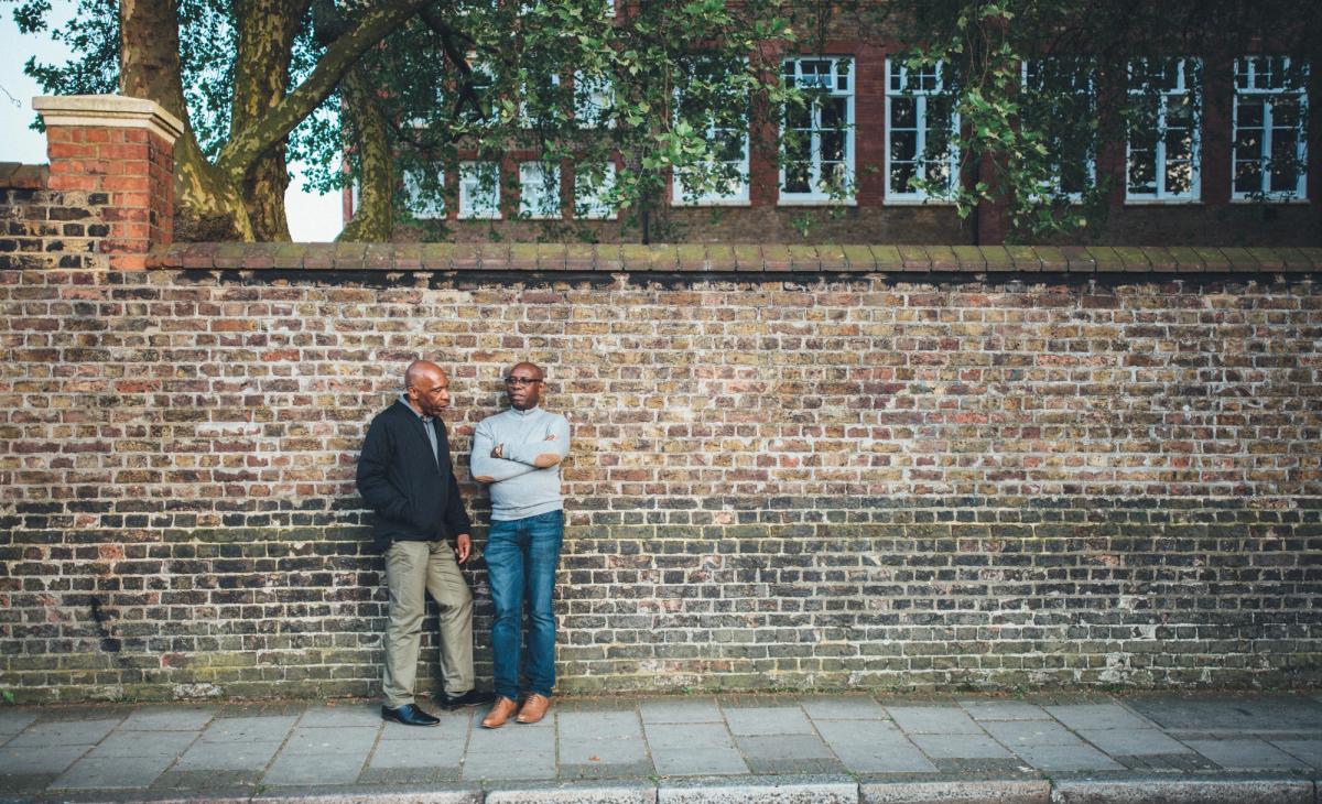 Two black men standing next to each other leaning on a brick wall outside