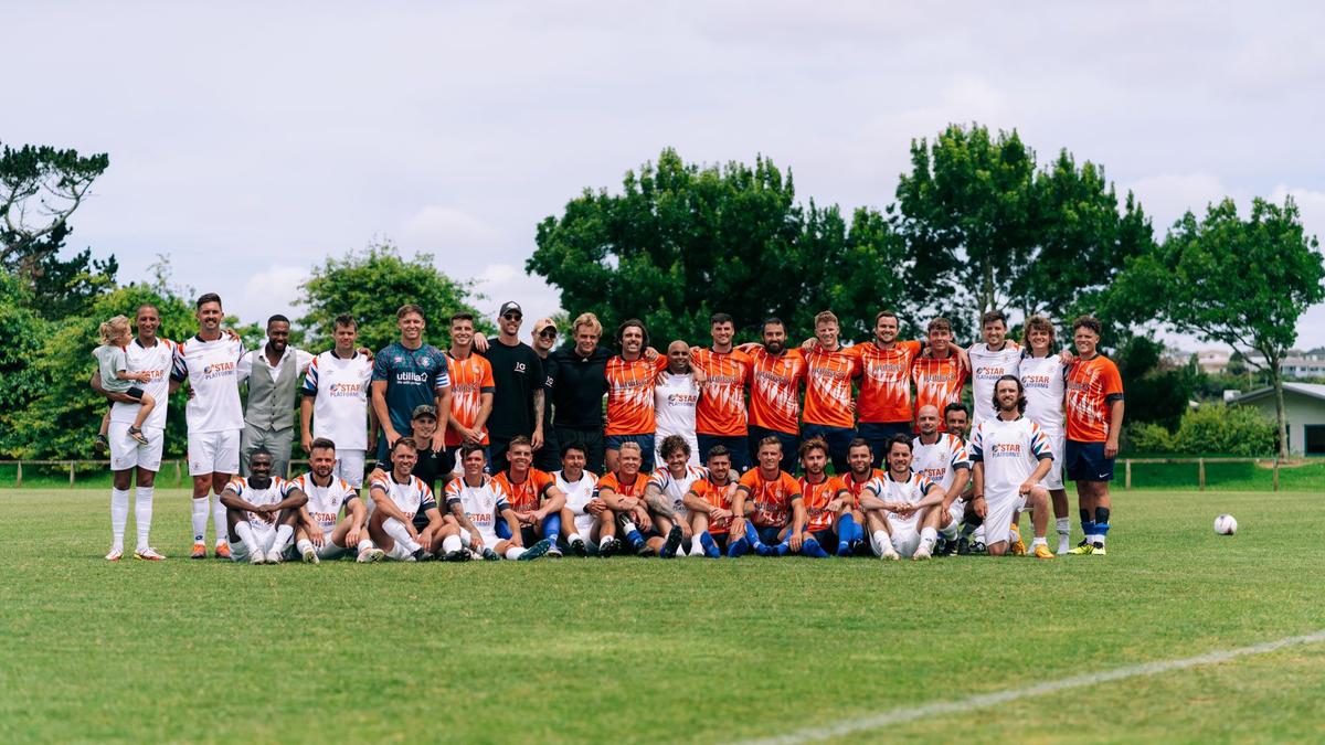 Two football teams take photo together on the grass pitch. Two lines, one sitting down in front and the other standing behind.