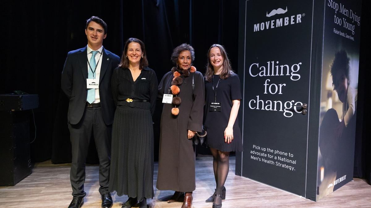 Four people pose in front of a Movember branded phone booth at a Movember event