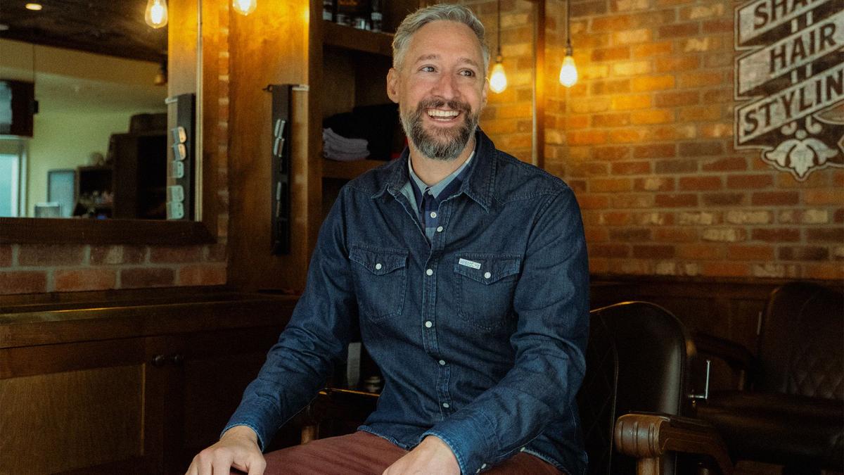 A man in a button up denim shirt smiles for a portrait in barber chair