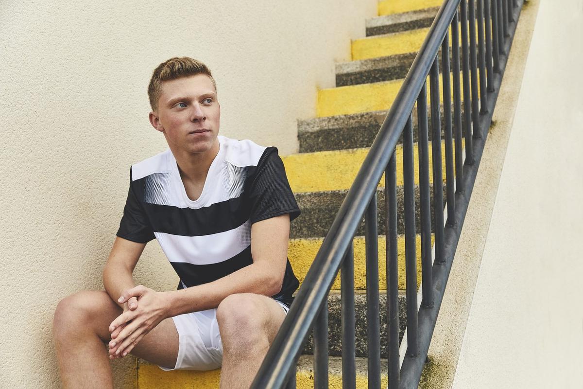 A young man in a sports kit sits on stadium stairs with hands clasped, gazing away from the camera in quiet thought.