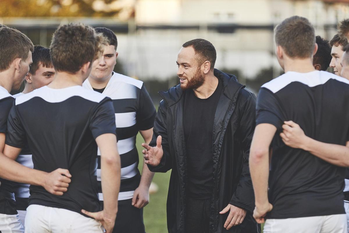 A coach in a black jacket talks to a group of young male rugby players huddled together on a field.