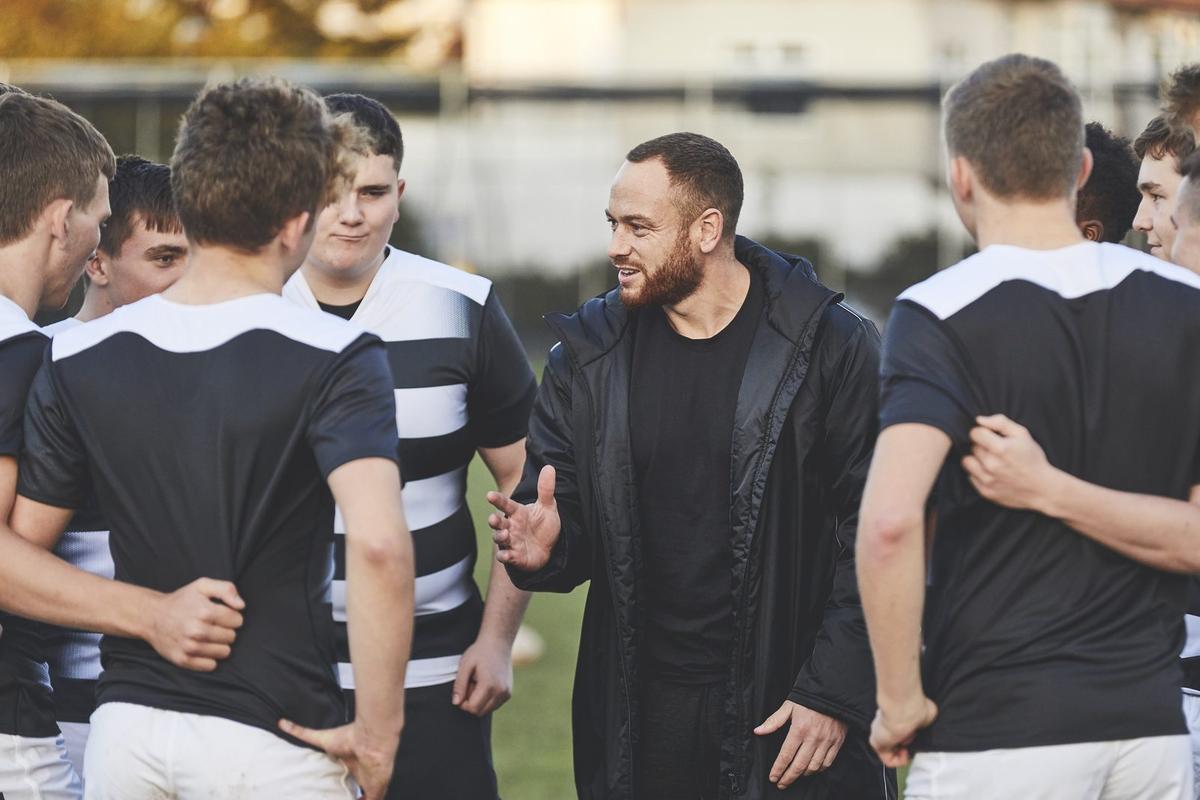 A coach in a black jacket talks to a group of young male rugby players huddled together on a field.