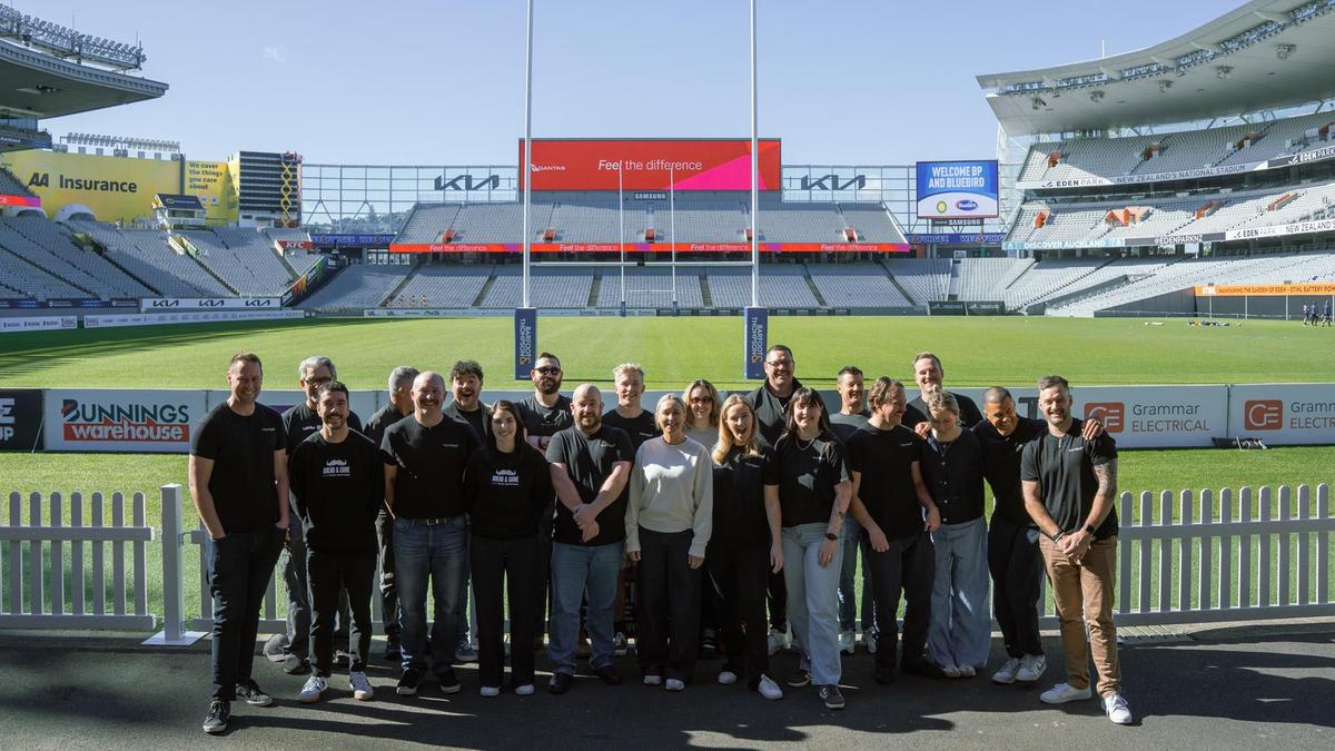 Group of people stand at eden park with goalpost in the background, sunny day