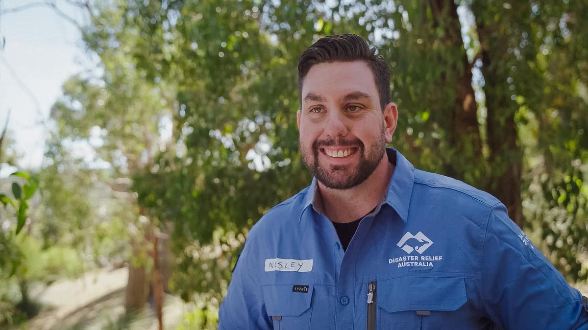 Man stands smiling at camera, tress in background, and blue sky above. He is wearing blue work overalls