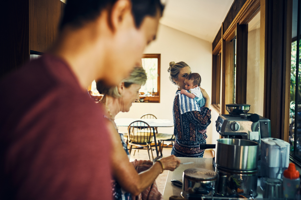 A mother is holding her baby in the kitchen. In the foreground, the father and grandmother are preparing a meal