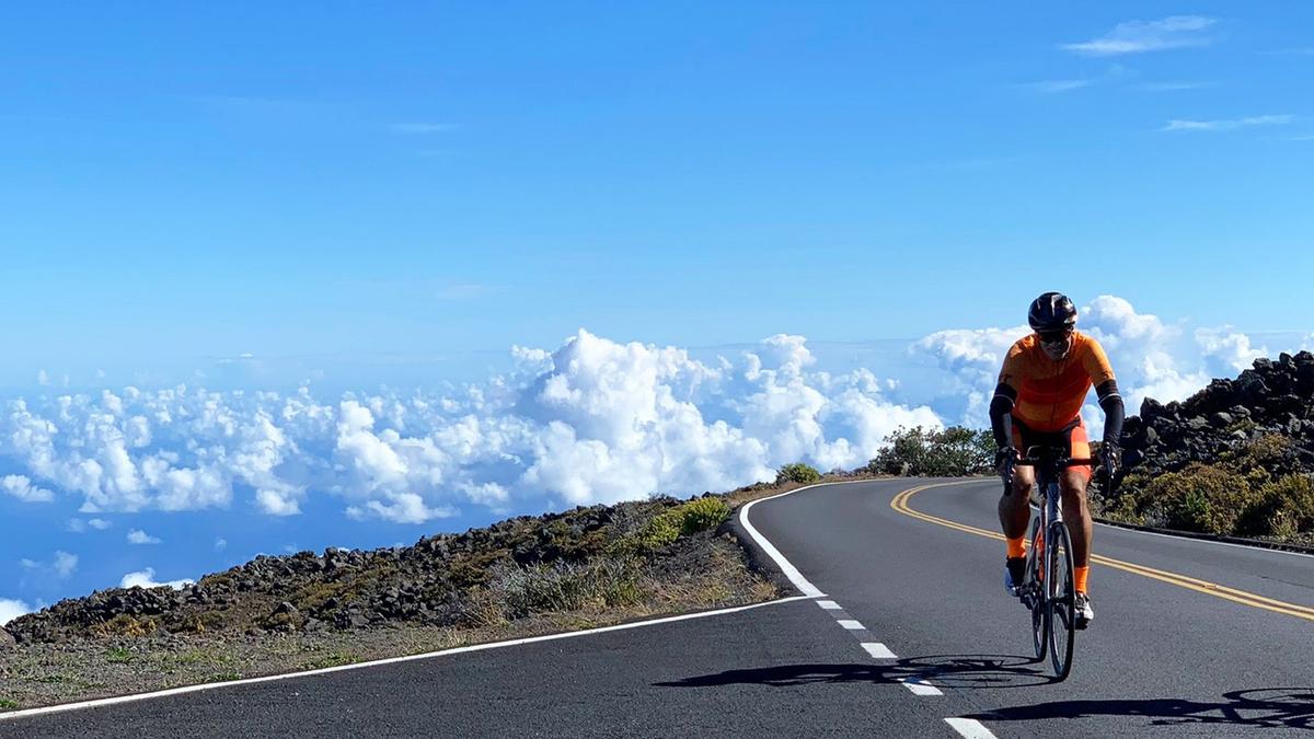 A man in an orange cycling suit cycles up a long road on Haleakalā Volcano in Maui. He's so high up that he's above the clouds.