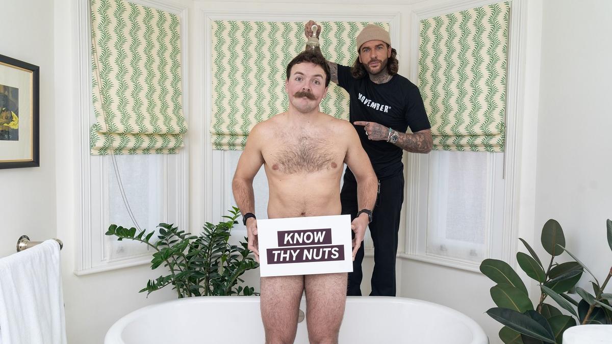 Pete holding a shower head over Sam in the bath, naked, holding a sign over his testicles saying 'know thy nuts'