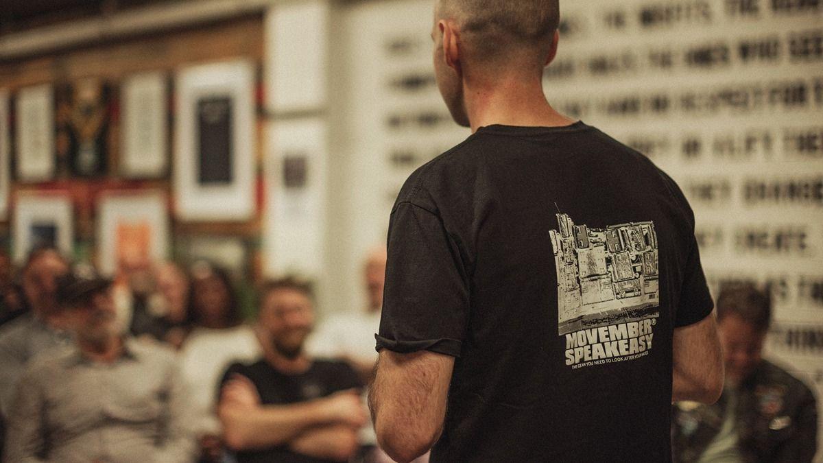 Man with back to camera speaking to a room of men