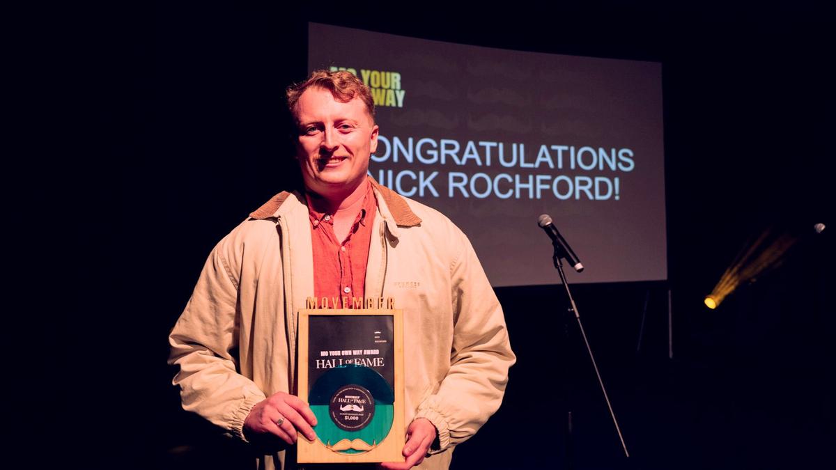Man holding Movember award on stage, mic to the left, background his name appears on screen.