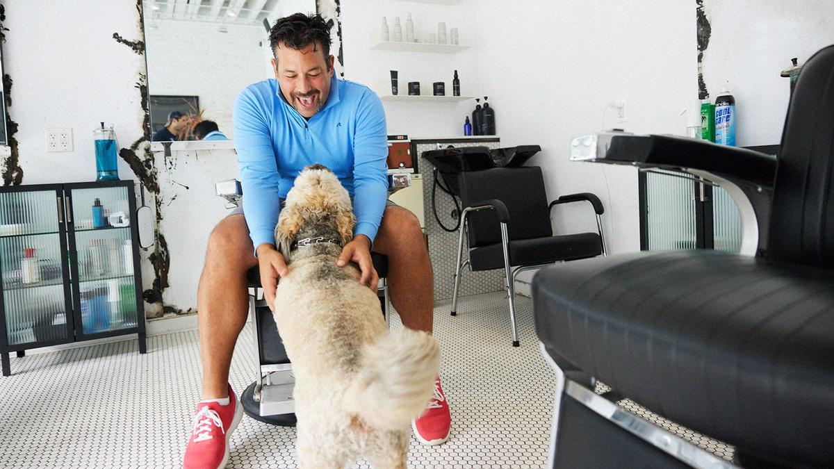 A man sits in a barberchair, excitedly smiling down and petting his dog who is wagging his tail from the ground