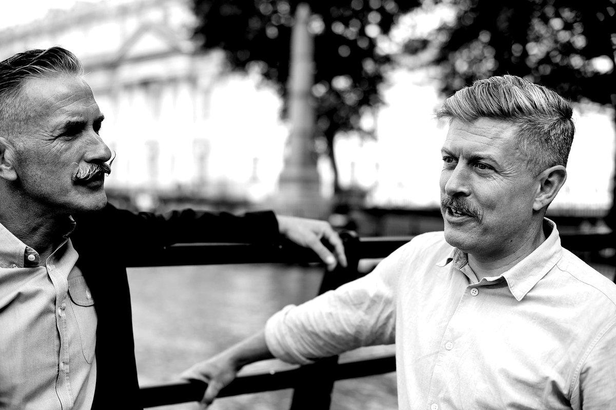 Two men with moustaches sit together outdoors, facing each other in conversation.