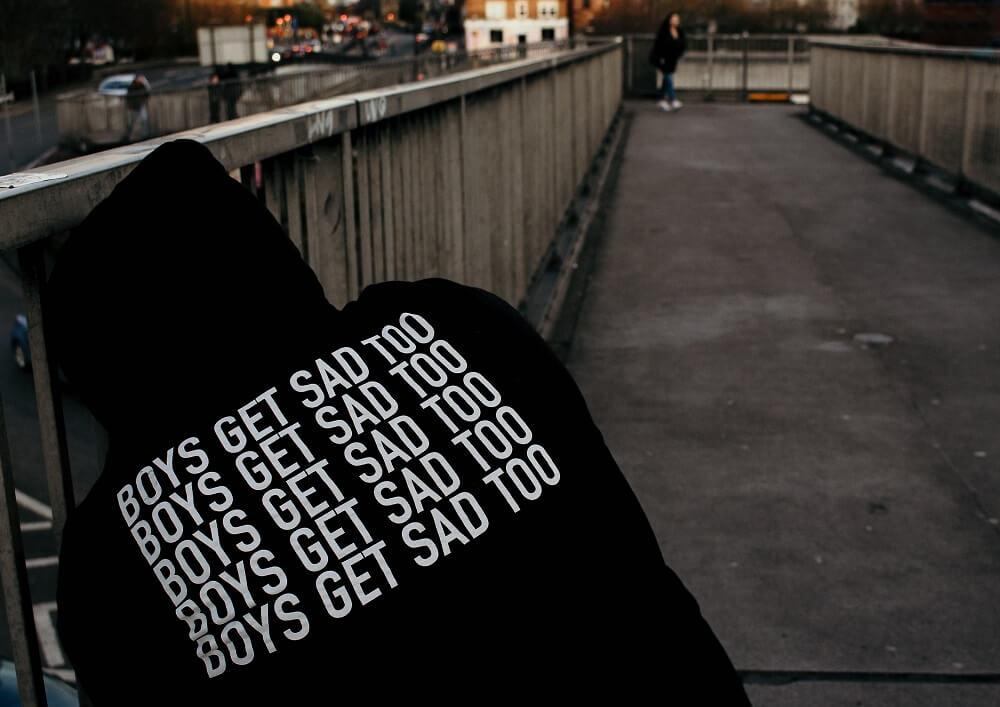 A black hoodie with "Boys Get Sad Too" printed on the back, on a bridge with a person walking in the distance.