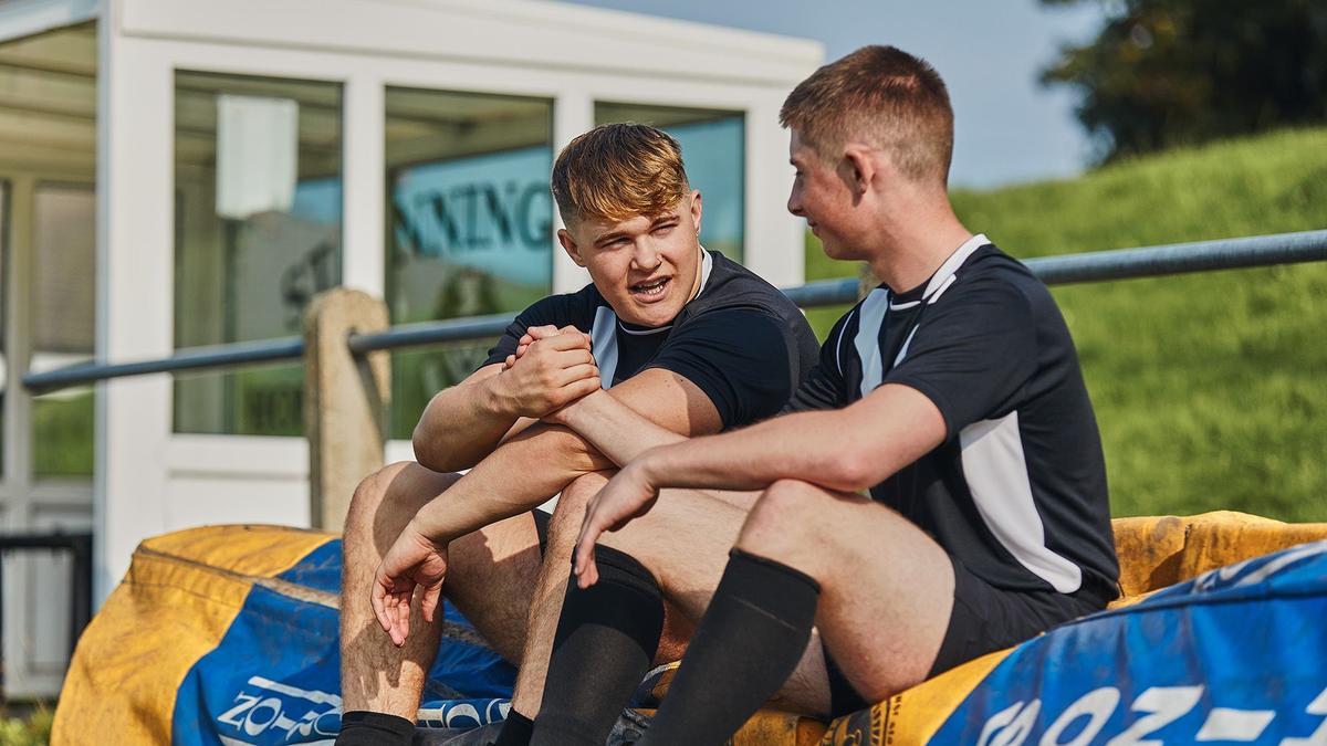 2 young men in sports attire sitting at a football ground and engaging with each other