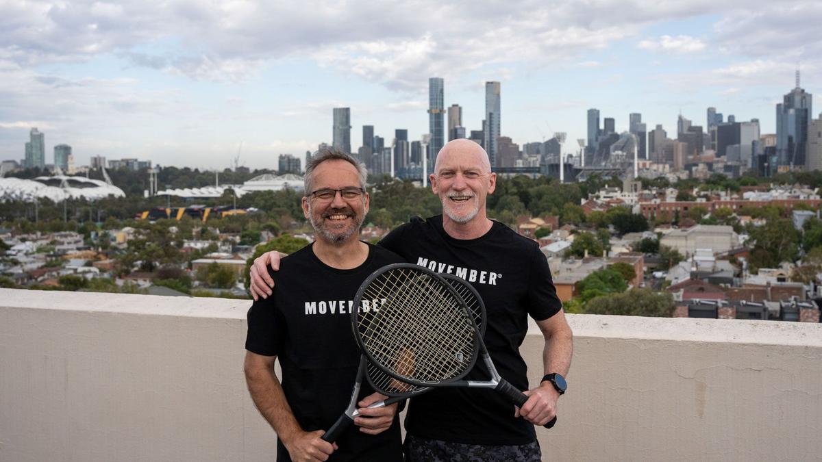 Two men holding tennis rackets, wearing Movember t-shirts, standing on rooftop with city scape in the background.