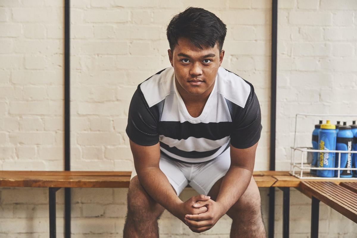 A young male rugby player sits alone in a locker room, leaning forward with hands clasped, looking directly at the camera.