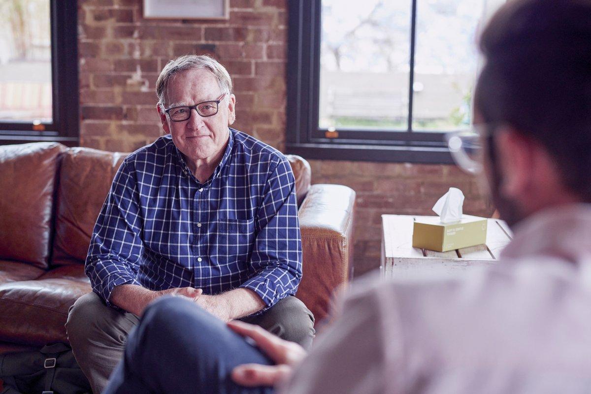 An older man in a blue checked shirt sits on a leather couch in a brick-walled room, smiling gently as he talks with another man seated across from him.
