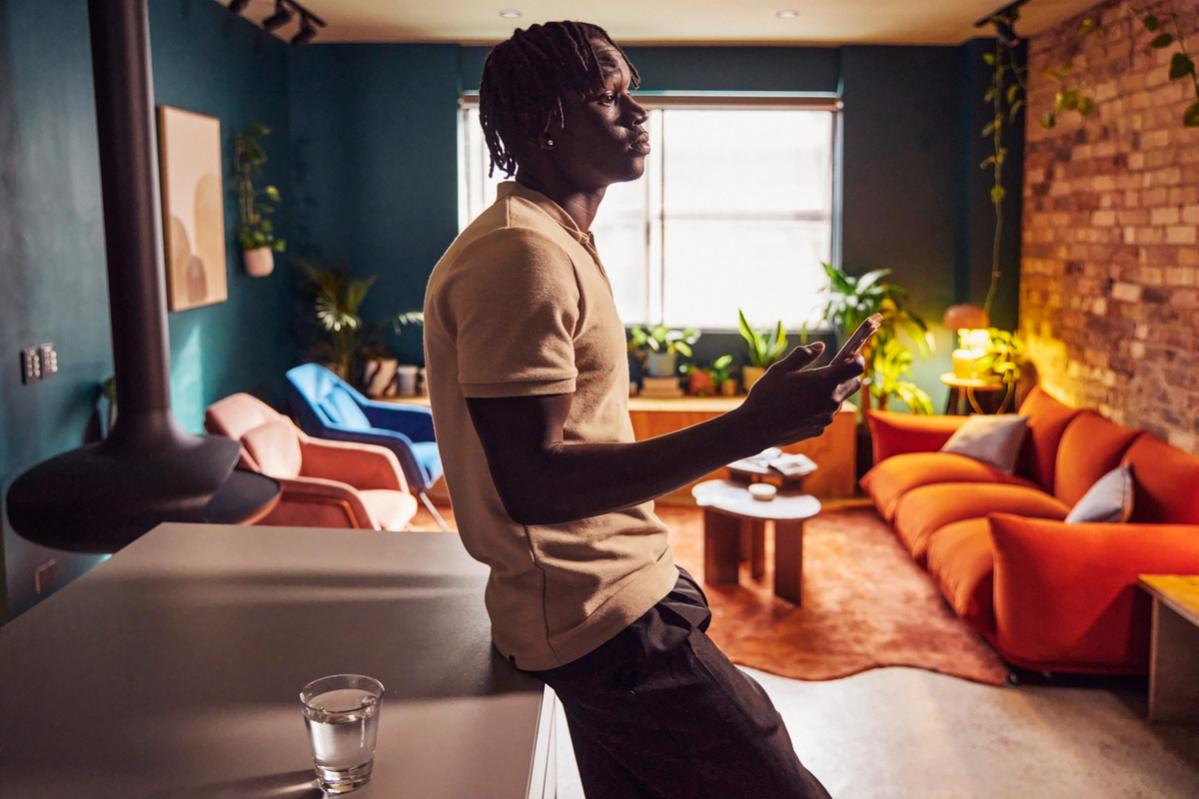 A black man slouching against a table holding his phone in an urban looking studio apartment