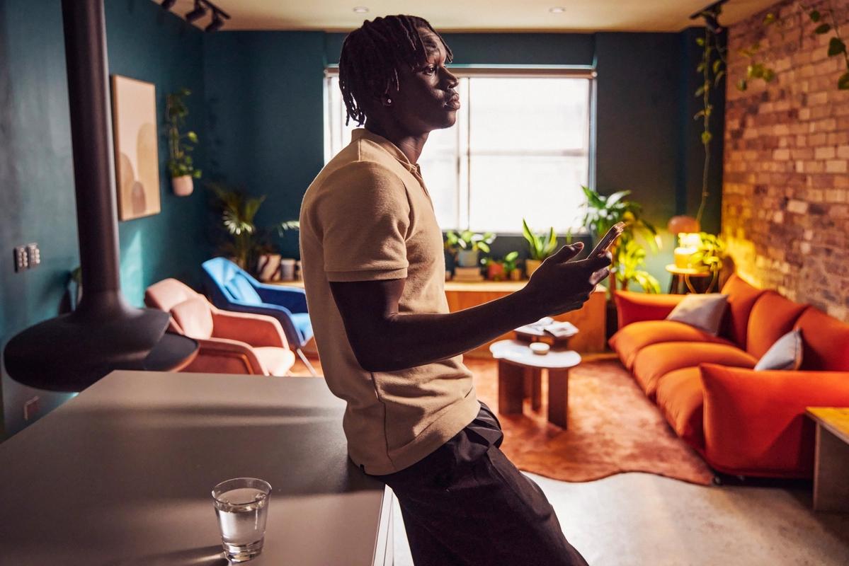 A black man slouching against a table holding his phone in an urban looking studio apartment