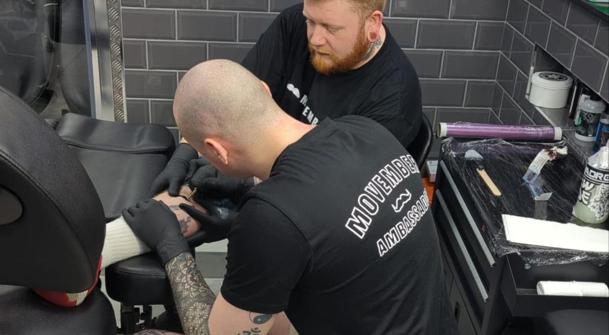 Man wearing a Movember t-shirt tattooing a moustache on another man's leg