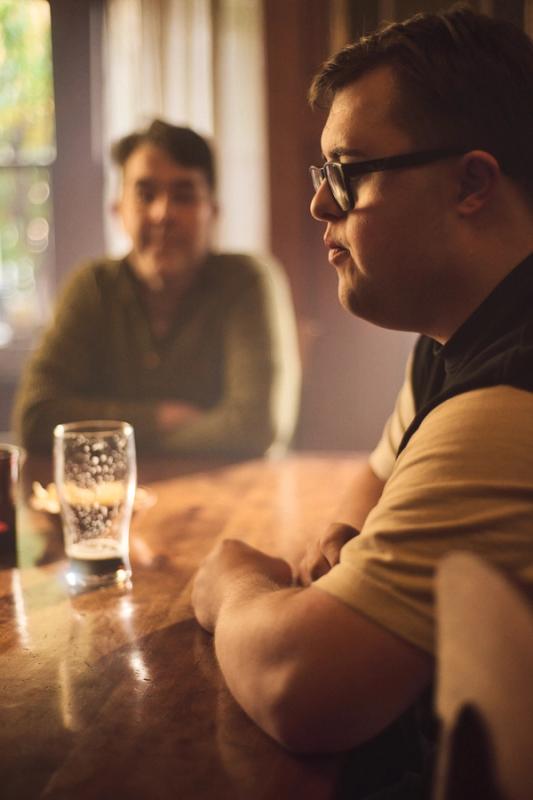 Two men are seated at a bar table in a pub, there's an empty pint of beer on the table