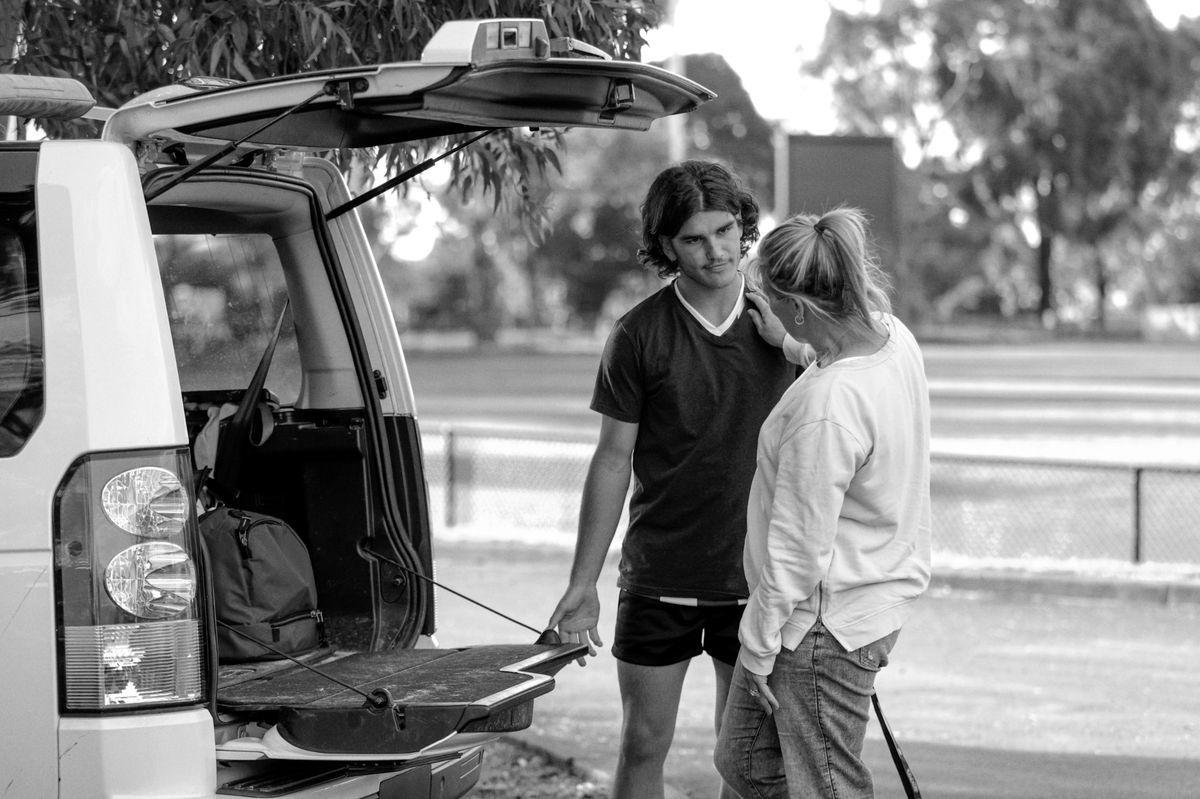 A mother and teenage son talk beside the open boot of a car.