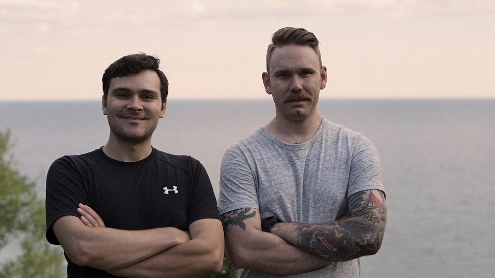 Two men stand together outdoors with the ocean behind them, arms crossed, looking at the camera.