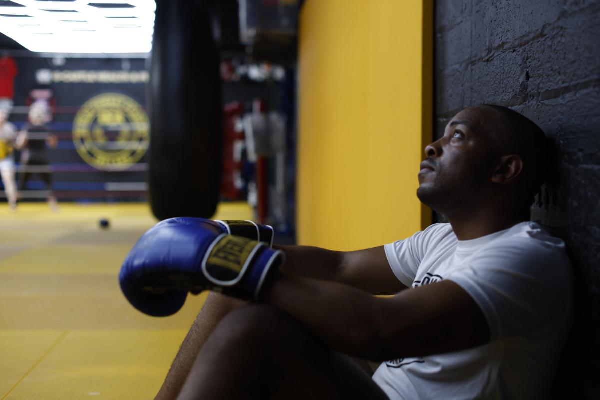 A black man sits on the floor with his back against the wall in the gym, he's wearing boxing gloves and looking thoughtful