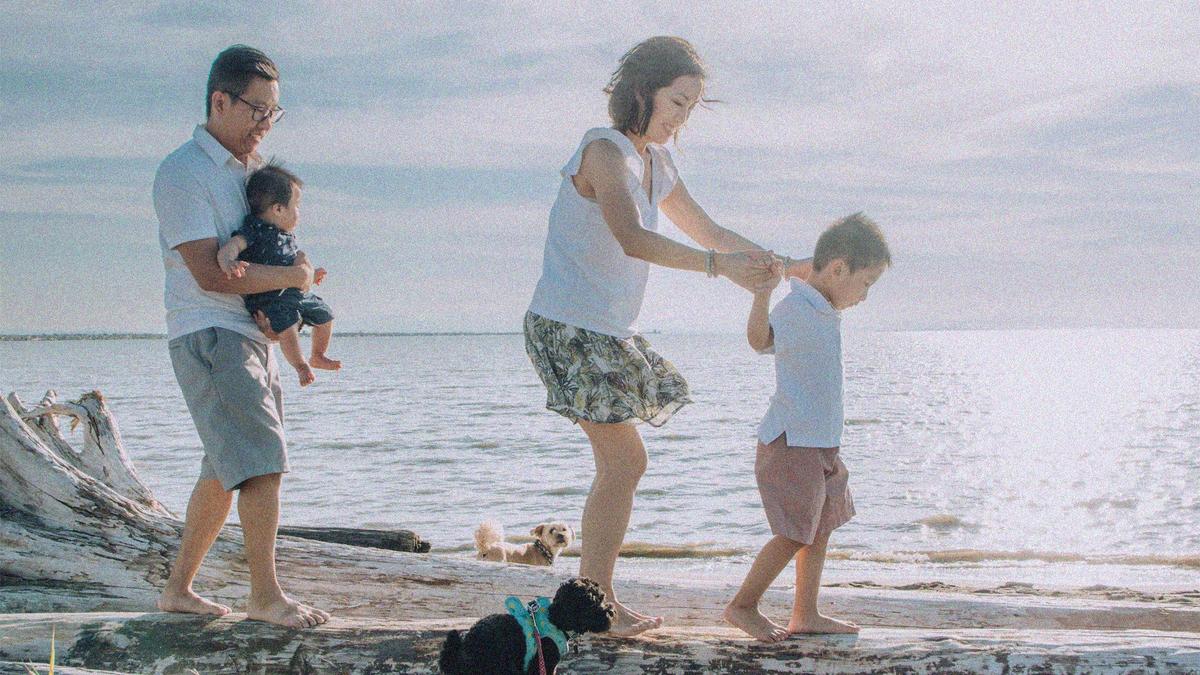 A man, holding his baby, walks on the beach with his wife, toddler, and 2 dogs